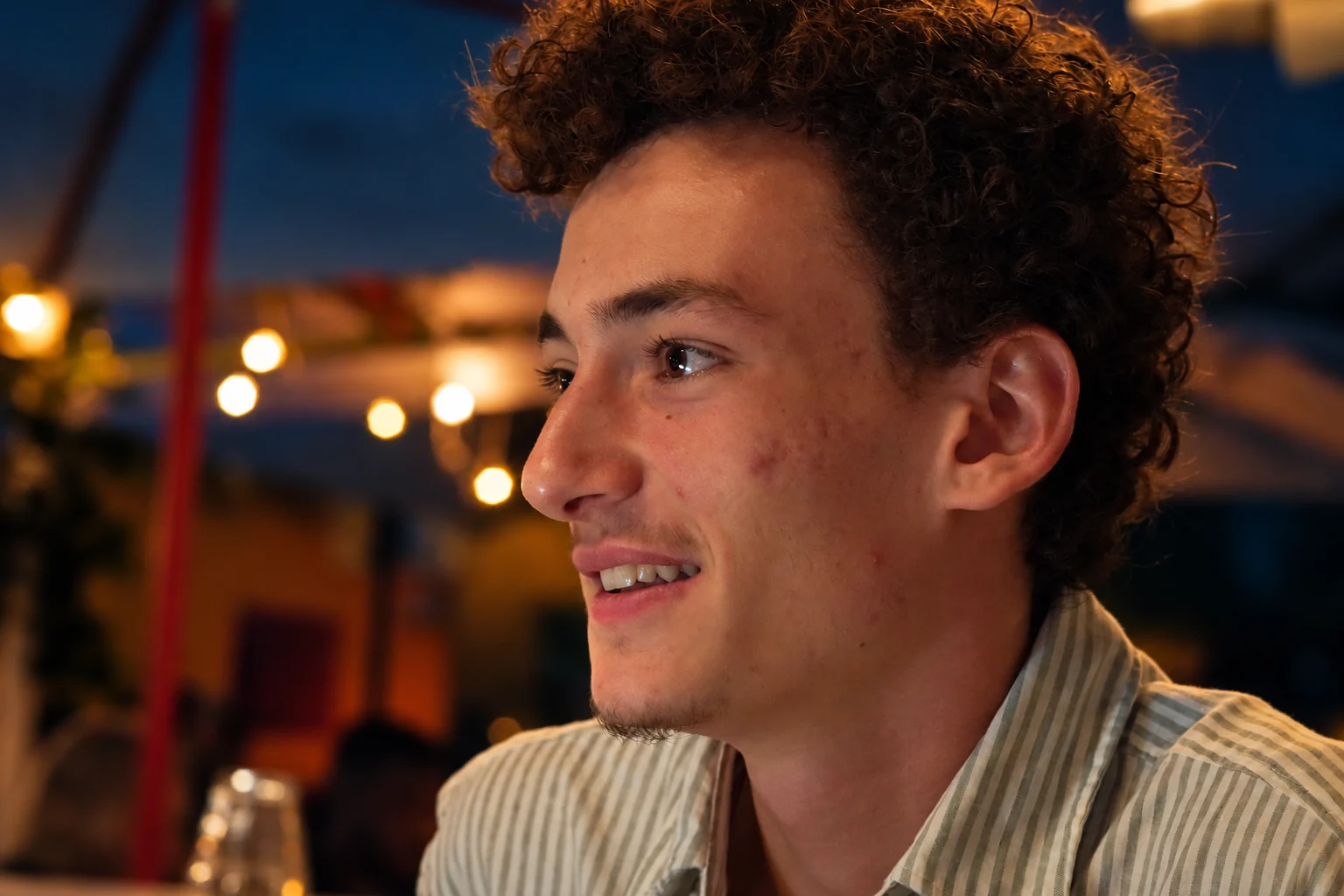 Un homme aux cheveux bouclés sourit de profil, entouré de lumières chaudes, série flo.